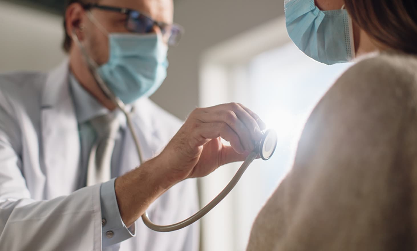 A doctor with thick frame glasses wearing a mask holding a stethoscope in front of a patient